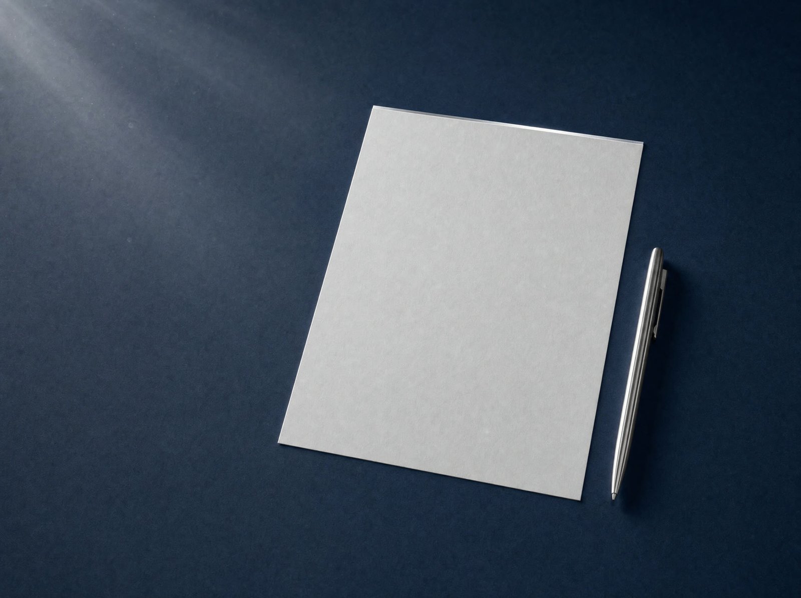 A blank sheet of heavyweight card beside a silver pen on a navy desk